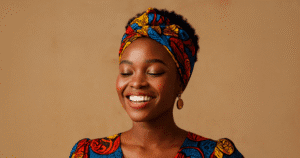 A close-up portrait of a young African woman with short, natural hair, wearing a vibrant, multicolored African print (Ankara) dress and matching head wrap. She has her eyes closed and is smiling broadly, showing a perfect, joyful expression. She is wearing round, amber-colored drop earrings against a plain tan background.